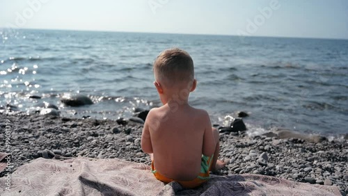 A boy-child is sunbathing, sitting with his back to the camera looking at the sea. Vacation on the seashore with the whole family. Slow motion video