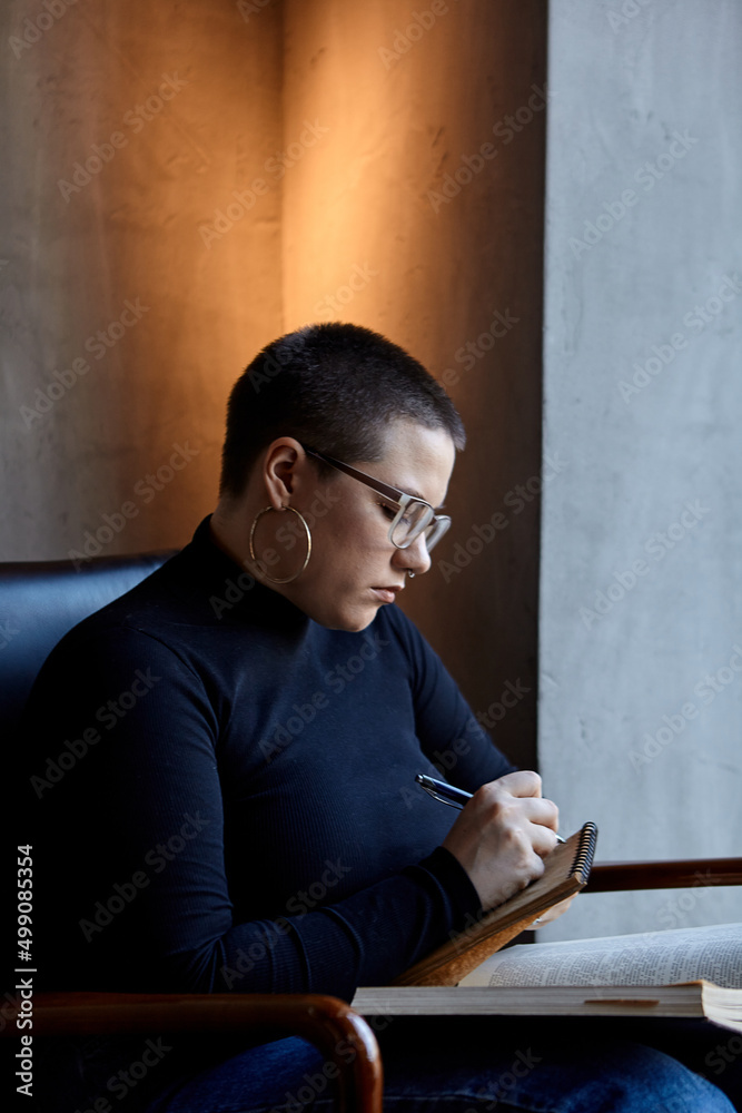 A young girl with glasses, with short hair is sitting in a cafe and reading a book. The girl looks at her book and makes notes in a notebook. The concept of distance learning A young girl with glasses, with short hair is sitting in a cafe and reading a book. The girl looks at her book and makes notes in a notebook. The concept of distance learning