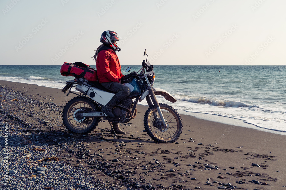 Woman riding enduro motorcycle standing on the shore near waves of the ocean. Female solo motorcycle trip