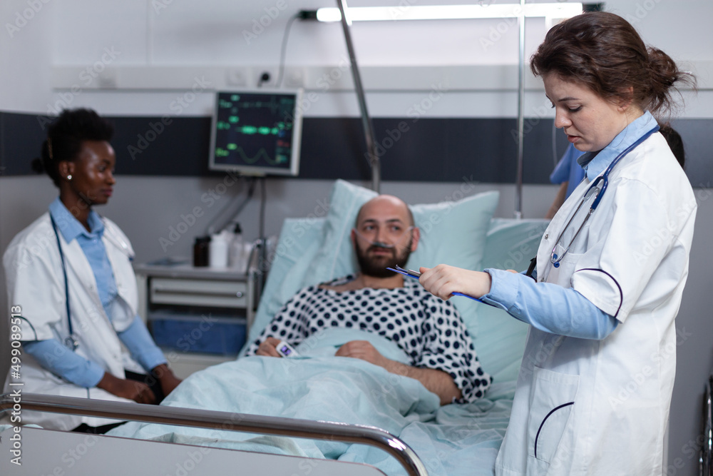 Fototapeta premium Middle aged man recovering lying in hospital bed attended by team of doctors after surgical intervention. Doctor with stethoscope reading medical chart while nurse is monitoring patient vitals.
