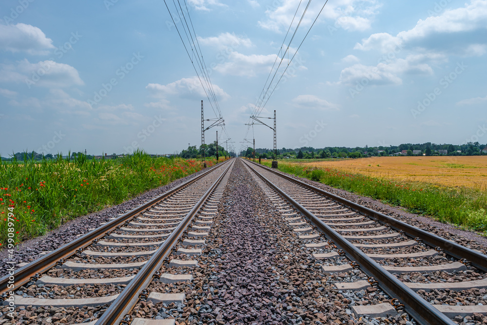 Railway for trains in beautiful farm landscape with wheat yellow field and poppy red flowers at warm sunset colors in summer, at sunny day and blue sky. Railway for trains in beautiful farm landscape with wheat yellow field and poppy red flowers at warm sunset colors in summer, at sunny day and blue sky.