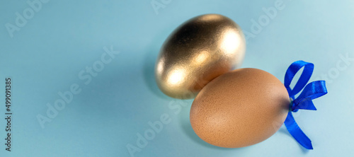 banner with Brown egg with blue ribbon and golden egg. Happy Easter concept. Two eggs with ribbon on a pastel blue background with copy space.