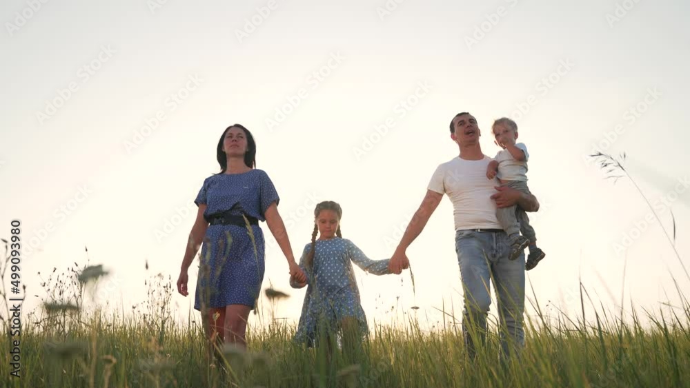 Happy family. Family walk in the meadow. Park with green grass. Parents hold their children by the hands in nature. Natural park at sunset. Active lifestyle. Cheerful happy family in the park outdoors Happy family. Family walk in the meadow. Park with green grass. Parents hold their children by the hands in nature. Natural park at sunset. Active lifestyle. Cheerful happy family in the park outdoors