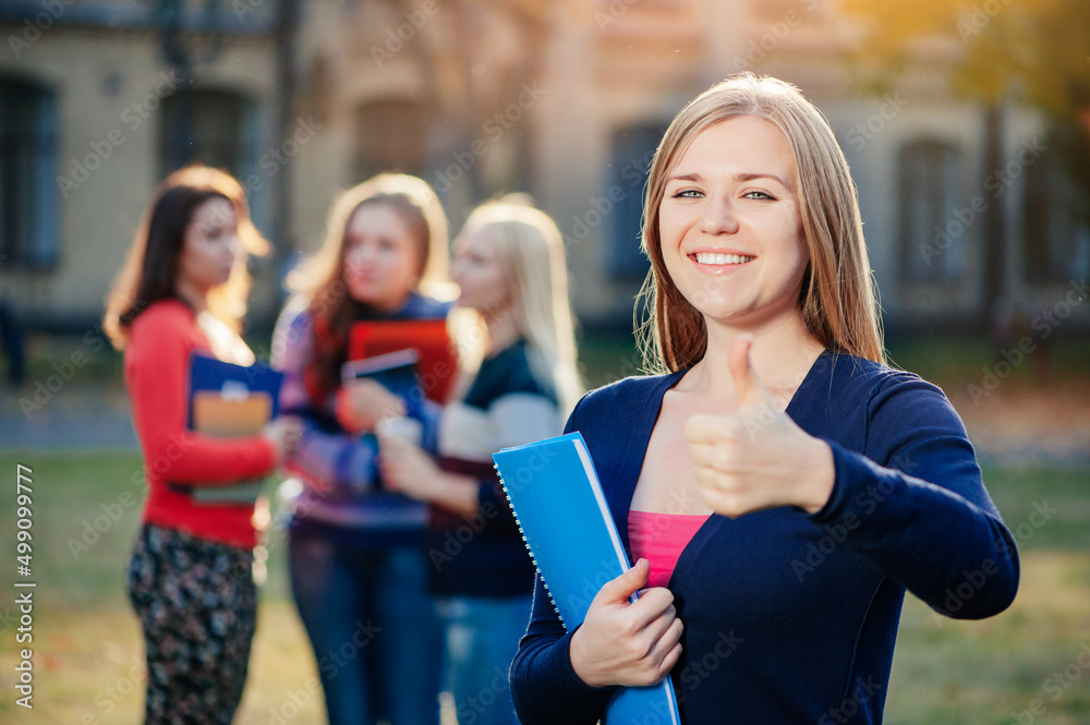 Thumb up for success! Handsome female student showing her thumb up and smiling while standing outdoors against university building while her friends communicating on background Thumb up for success! Handsome female student showing her thumb up and smiling while standing outdoors against university building while her friends communicating on background