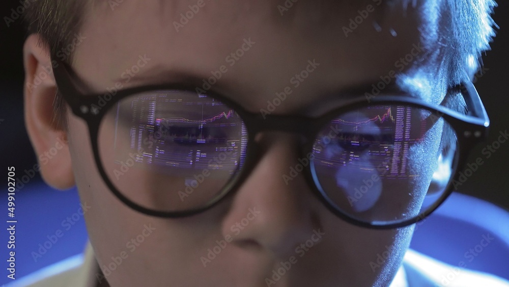 Close-up of the face of a boy with glasses sitting at a computer and looking at an online stock market chart showing bitcoin currencies. The glasses reflect the bitcoin graph. In real time Close-up of the face of a boy with glasses sitting at a computer and looking at an online stock market chart showing bitcoin currencies. The glasses reflect the bitcoin graph. In real time