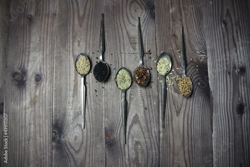 Metal spoons with spice seeds of anis, nigella, ajowan, coriander, rosemary, Sichuan pepper on brown wooden background