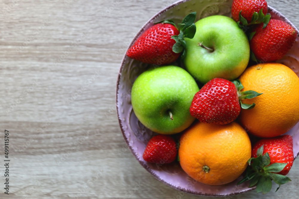 Pink bowl filled with fresh apples, oranges and strawberries on wooden table. Flat lay.