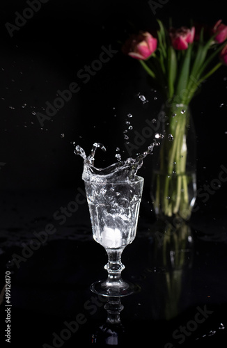 Glass of splashing water on a black background with pink tulips on the back