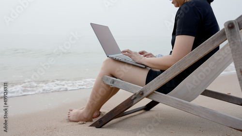 Close-up of a person sitting on a chair by the sea, where the sea waves are slowly hitting the shore, and hands are typing some information on a laptop computer.