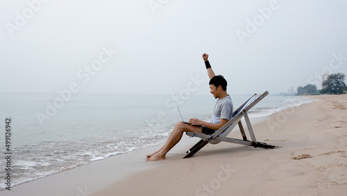 A handsome Asian man sits on a chair in a laptop computer by the sea as the waves crash against the shore, he raises his hand glad to offer the job, pleasantly by the sea, work from home.