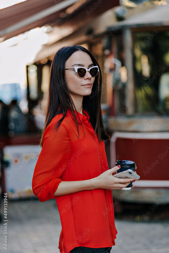 Obraz premium Elegant attractive european woman walking outdoors with coffee. Woman in sunglasses wearing red blouse while listening to music.