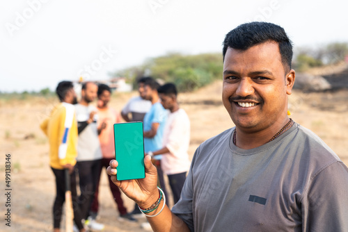 Smiling cricket player showing mobile phone by looking camera at playground in front of players - concept of online booking, app promotion and advertisement