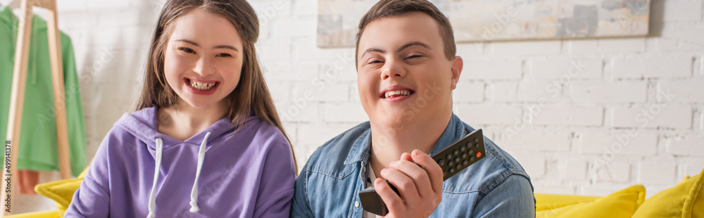 Positive teenagers with down syndrome holding remote controller at home, banner. Positive teenagers with down syndrome holding remote controller at home, banner.