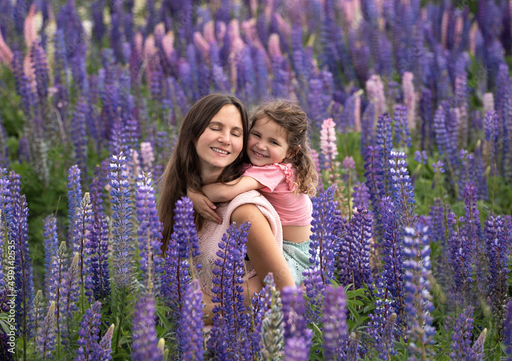 Young mother looking at her little daughter with curly blond hair and rose summer dress in lupine flower field. Young mother looking at her little daughter with curly blond hair and rose summer dress in lupine flower field.