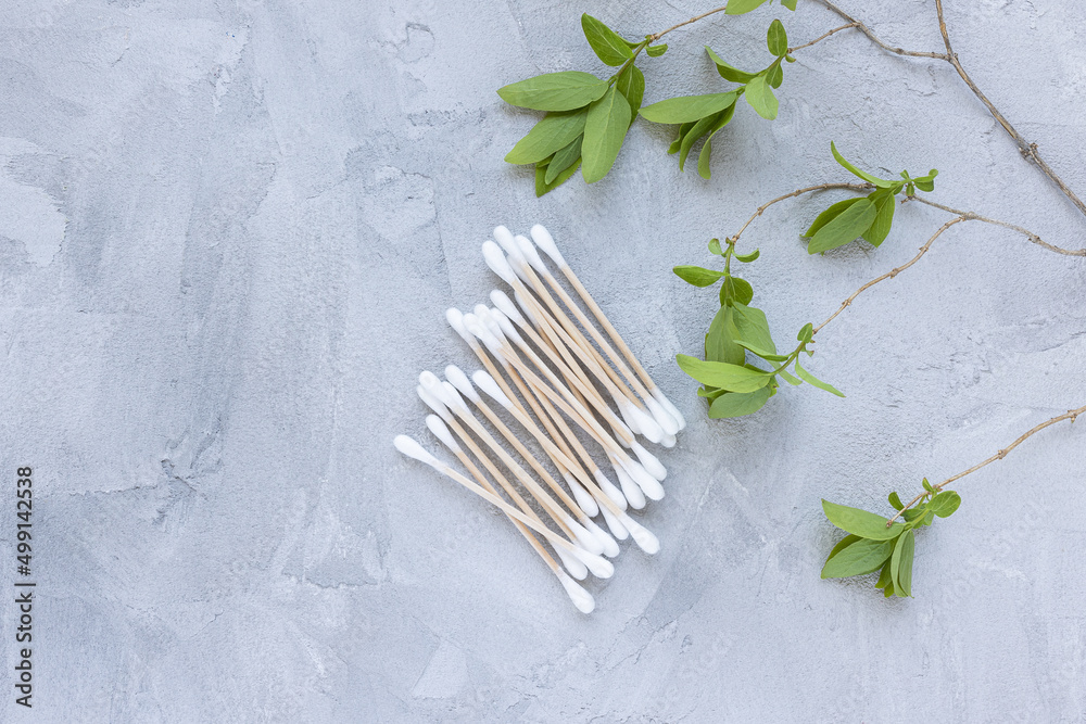 Eco-friendly bamboo cotton buds on gray background with green leaves. Trendy cotton swabs, bamboo ear cleaner sticks. Flat lay, top view, copy space