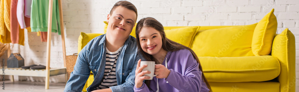 Smiling teen girl holding cup and looking at camera near boyfriend at home, banner. Smiling teen girl holding cup and looking at camera near boyfriend at home, banner.