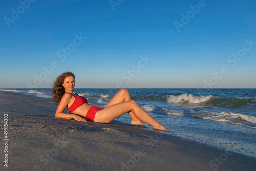 Portrait of sexy woman in red swimsuit. Girl lies on empty sea shore before sunset