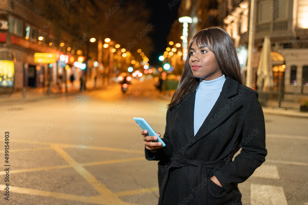 Fototapeta premium Young woman standing on street holding her phone in city night