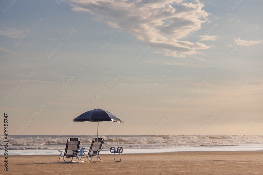 Conjunto com duas cadeiras de praia, um carrinho de transporte e um guarda-sol, sobre a areia da praia vazia e um lindo entardecer. 