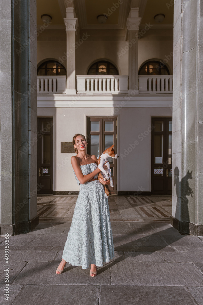 Obraz premium An incredibly beautiful woman with a dog laughs. She is dressed in a gently turquoise taffeta dress. Luxurious hairstyle and red lipstick make woman bright and memorable