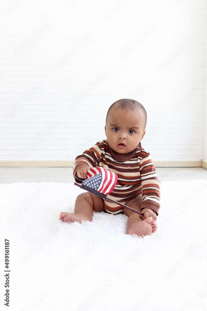 African American infant baby sitting on floor in indoor living room, little kid holding and waving American flag on 4th July to celebrate national Independence Day, Flag Day concept. African American infant baby sitting on floor in indoor living room, little kid holding and waving American flag on 4th July to celebrate national Independence Day, Flag Day concept.