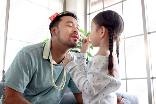 Joyful father playing with his little adorable daughter together, girl doing makeup to her dad by cosmetic, sitting on sofa in living room, have fun and standing time on family holiday weekend.