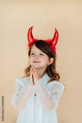 Portrait of a little girl in a white dress with devil horns. Halloween costume. Angelic facial expression. The concept of two different personalities in character.
