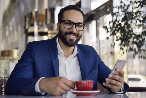 Handsome smiling Arabian businessman using mobile phone drinking coffee in cafe. Portrait of  middle eastern man wearing eyeglasses holding smartphone working online in office. Successful business 
