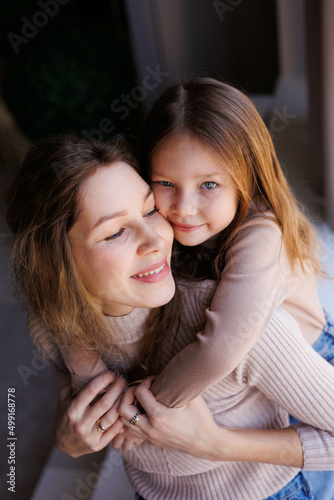 Lovely family mother and daughter embracing and kissing each other indoors. Happy sweet woman with little 6-year-old girl in casual clothers: turtlenecks and jeans sitting on sofa, hugging and smiling