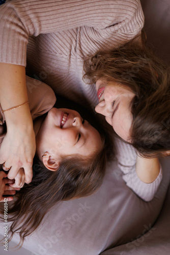 Lovely family mother and daughter embracing and kissing each other indoors. Happy sweet woman with little 6-year-old girl in casual clothers: turtlenecks and jeans sitting on sofa, hugging and smiling photo