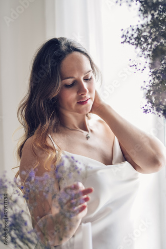Obraz na płótnie Portrait of young beautiful long-haired woman in a light silk dress at the window in the room decorated with gypsophila trendy color of 2022 year very peri. w salonie