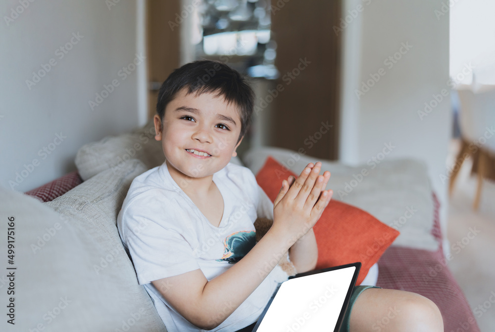 Happy young boy looking at camera with smiling face and clapping hands, Positive kid using tablet playing game online at home, Child sitting on sofa relaxing on weekend Happy young boy looking at camera with smiling face and clapping hands, Positive kid using tablet playing game online at home, Child sitting on sofa relaxing on weekend