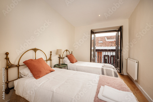 Bedroom with two single beds with brass headboards  circular wooden table  white aluminum radiator  balcony overlooking the roofs of vintage buildings