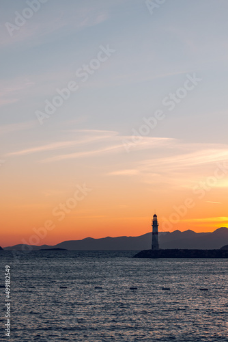 A white lighthouse with a red stripe on the pier. Sunset in the bay of the Aegean Sea among the mountains. Greece, Bodrum, Turkey.