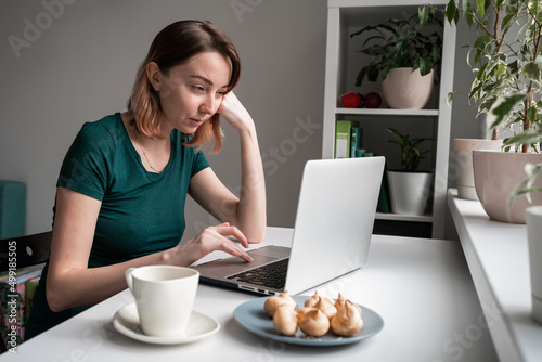 Young woman sitting by the window with laptop. Tea or coffee with sweets.