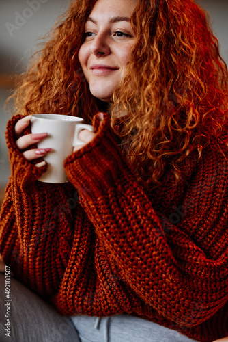 A cute young woman with red curly hair is enjoying her cup of coffee in the morning.