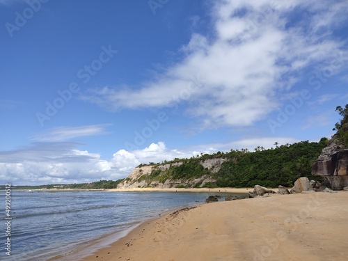 The sea, the beach, sand, blue sky, trees and rocks in the background.