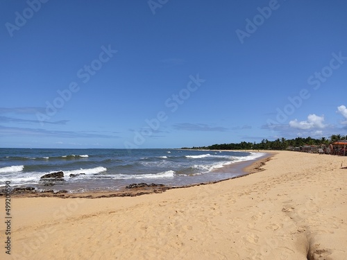 The sea  the beach  sand  blue sky  trees and rocks in the background.