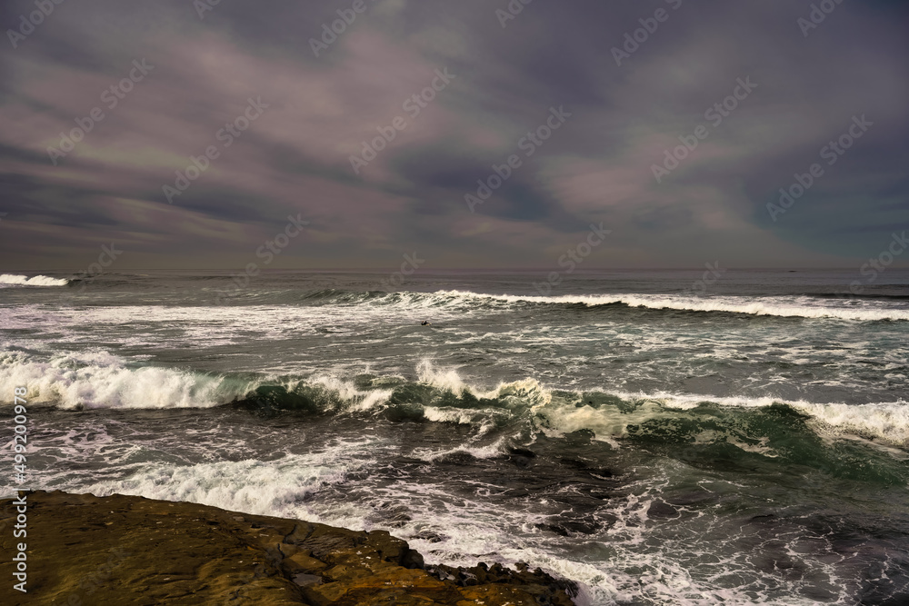 2022-04-15 WAVES CRASHING ON THE SHORELINE IN LA JOLLA CALIFORNIA WITH A LONE SURFER IN THE SEA AND A SOFT LIGHT TONED BLURRY SKY