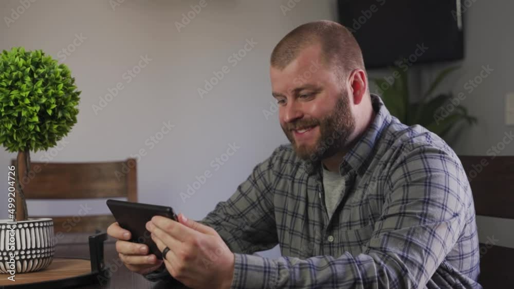 A Friendly Looking Bearded Young Adult Waving Hand While Video Chatting With Someone On His Tablet At Home. - Medium Closeup Shot A Friendly Looking Bearded Young Adult Waving Hand While Video Chatting With Someone On His Tablet At Home. - Medium Closeup Shot