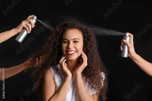 Beautiful young African-American woman and hands with hair spray on black background © Pixel-Shot