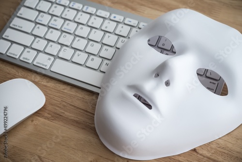 White scary half face mask, keyboard and mouse on wooden table background in dark tone. Hacker, cybercrime, romance scam and online security concept. photo