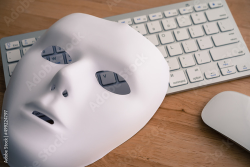 White scary half face mask, keyboard and mouse on wooden table background in dark tone. Hacker, cybercrime, romance scam and online security concept. photo
