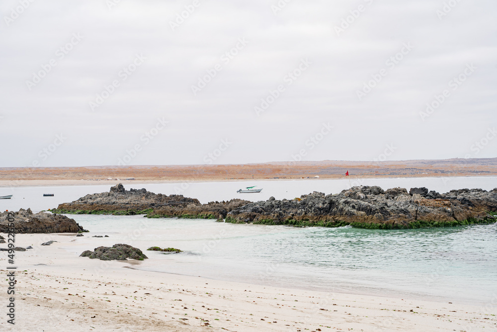 Horizontal view of turquoise beach on a cloudy day with rocks, boats and desert in the background.