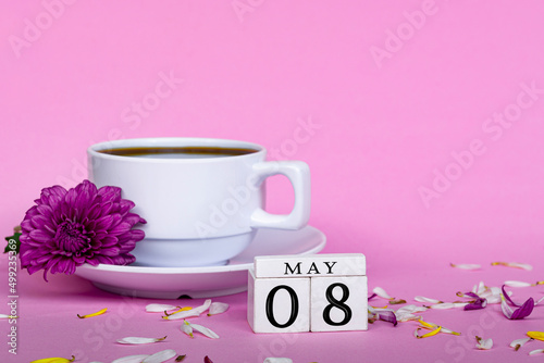 White coffee cup and purple flower with wooden block cube on pink background.