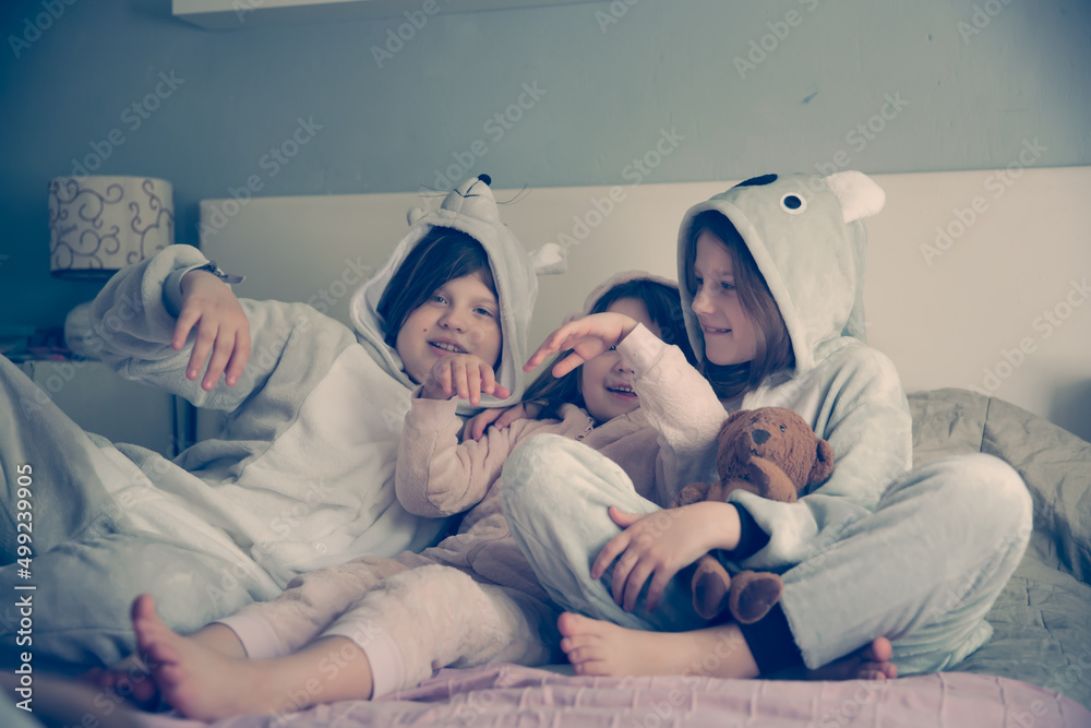 Four girls girlfriends sisters in pajamas at home on the bed, a pajama party in soft amigurumi costumes in a light gray bedroom, four children girls hugging on the bed Four girls girlfriends sisters in pajamas at home on the bed, a pajama party in soft amigurumi costumes in a light gray bedroom, four children girls hugging on the bed
