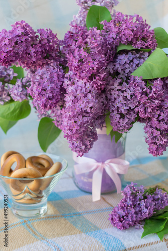 Vertical photo of a bouquet of flowers with lilac petals in a vase with a bow on a table with a checkered tablecloth and a vase with sushki