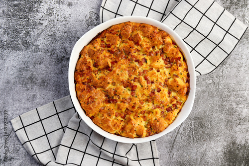 Breakfast Strata casserole with ham, herbs, cheese and cream in a white baking dish on a dark grey background. Top view, flat lay.
