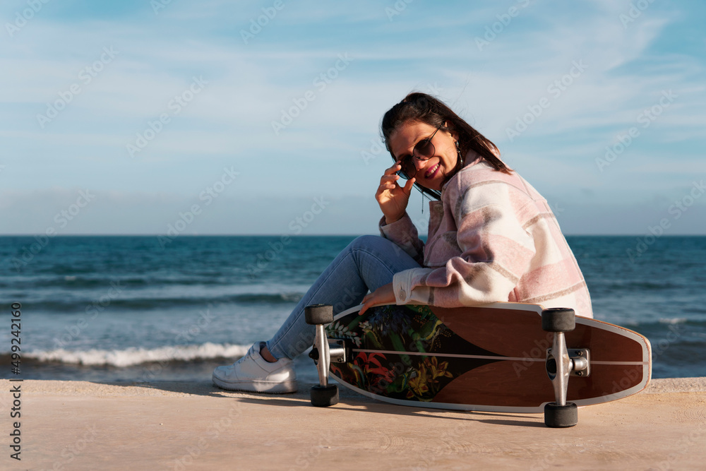 Woman wearing sunglasses, shirt and jeans sitting on the boardwalk next to her longboard. Young girl sitting sideways leaning on her skateboard with the sun on her back and fresh air.