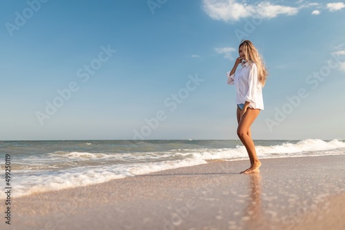 A slender girl in a gentle blue swimsuit and shirt walks along the sandy beach near the blue sea